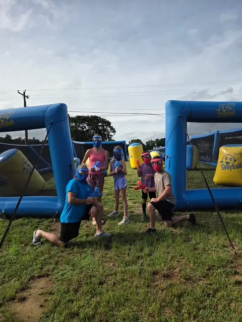 Group of kids and adults in blue masks pose with GellyBall guns beside inflatable blue arena barriers on grass.