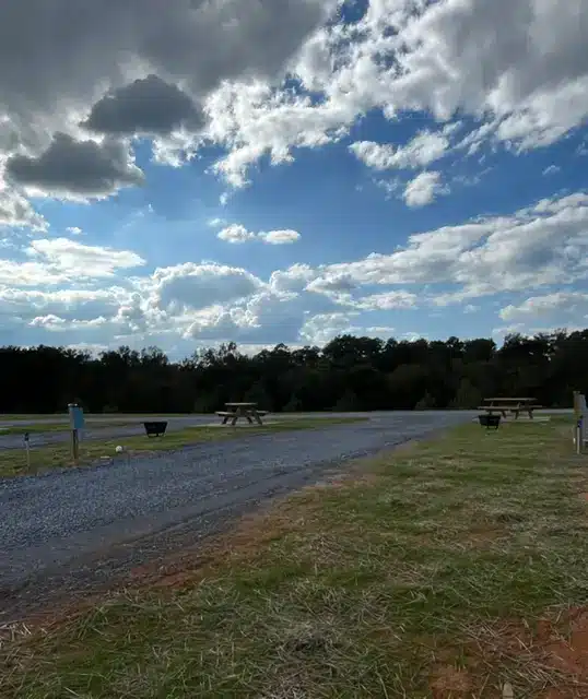 Empty gravel RV sites with wooden picnic tables and black fire rings beside a grassy field under dramatic cloudy sky.