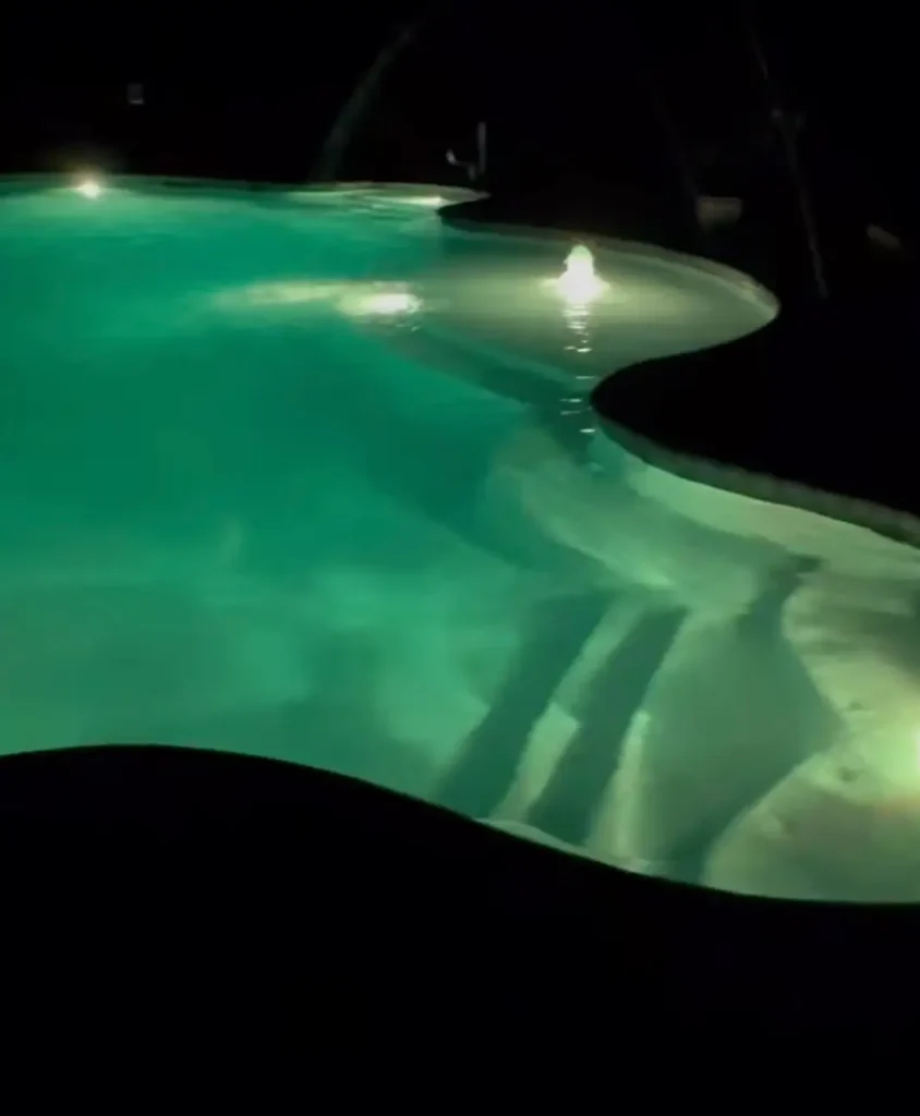 Nighttime photo of a curved freeform swimming pool glowing green from underwater lights, with dark surroundings and