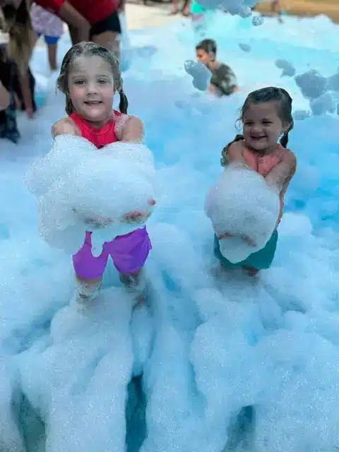 Two smiling young girls hold armfuls of white foam during an outdoor foam party, with other kids playing behind them.