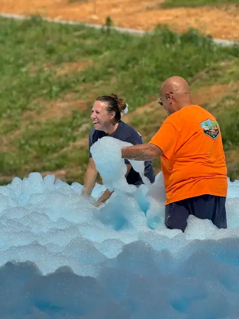 A smiling woman and a man in an orange shirt wade through a large blue foam party pit outdoors on grass.