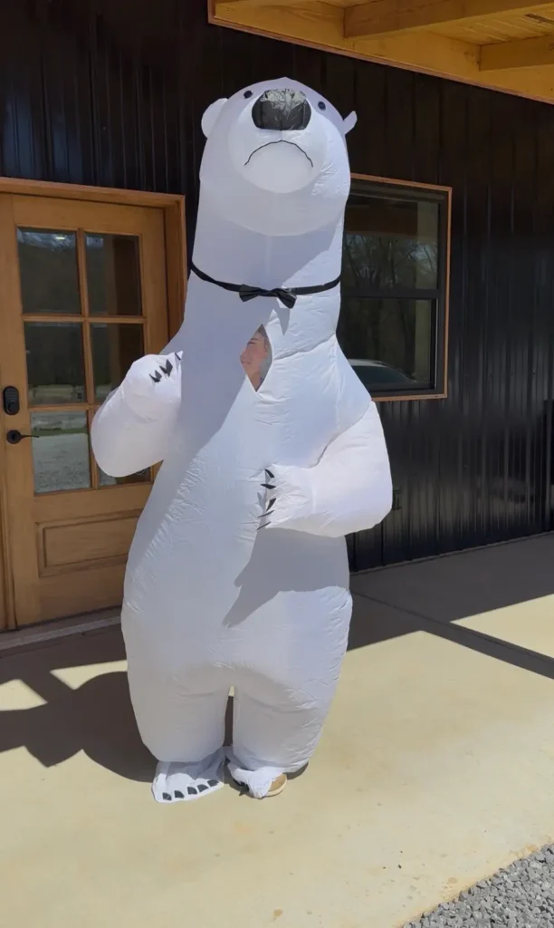 Person wearing a large inflatable white polar bear costume standing on a concrete patio outside a dark metal building