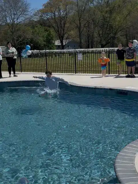 A person splashes into a clear outdoor swimming pool while children and adults watch from the fenced deck with balloons.