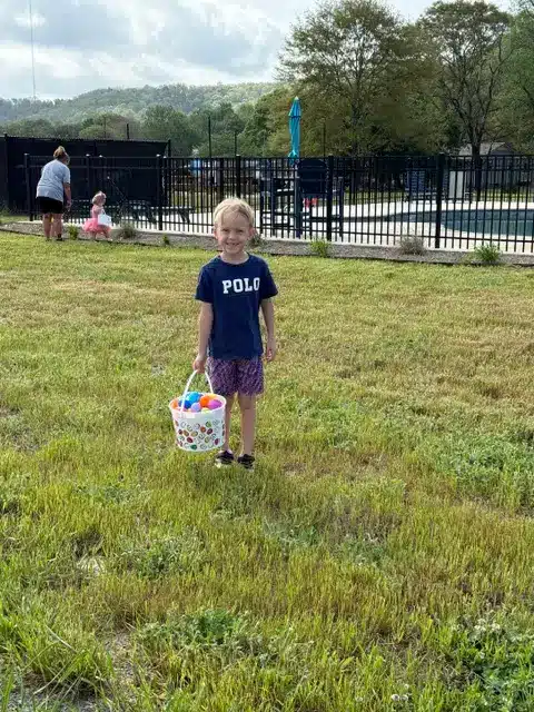 Young boy in a navy POLO shirt holds a bucket of colorful Easter eggs on a grassy field near a fenced pool area.