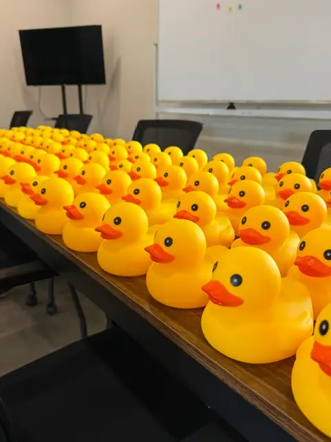 Dozens of yellow rubber ducks lined up on a wooden conference table in a meeting room with whiteboard and TV.