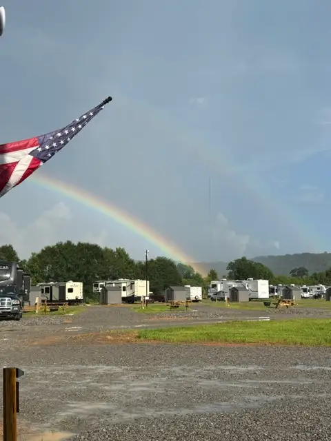 A double rainbow arcs over a gravel RV park with parked trailers, picnic tables, trees, and an American flag.