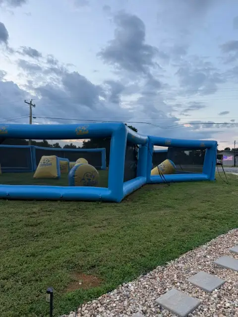 Large blue inflatable archery tag or paintball arena with yellow bunkers set up on grass at dusk.