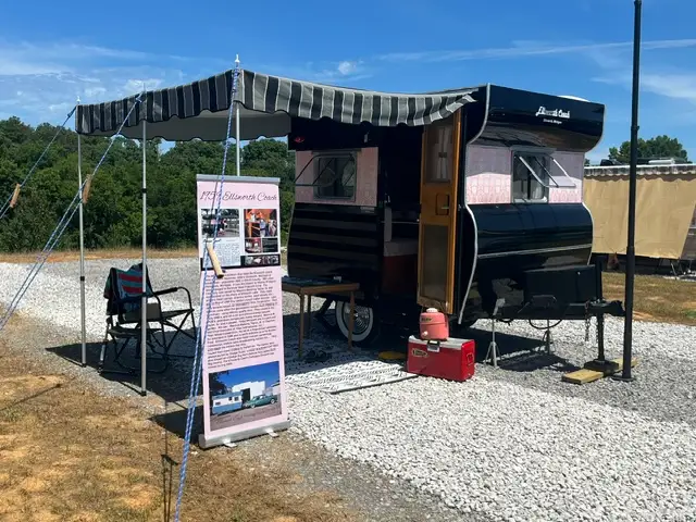 Vintage black camper trailer displayed on gravel lot with striped awning, folding camp chairs, red cooler, and