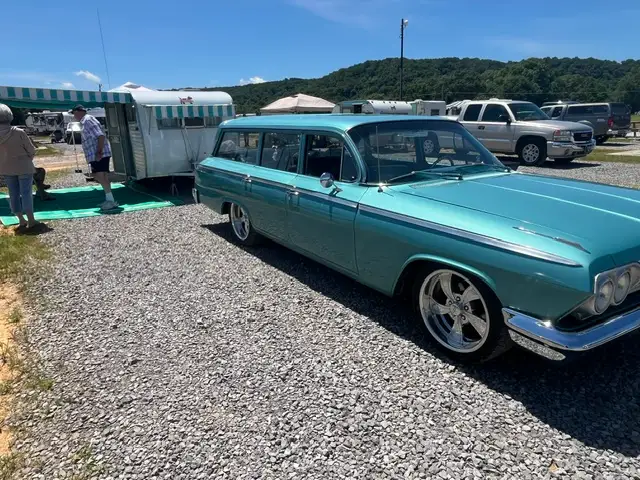 Restored turquoise 1962 Chevrolet station wagon parked on gravel beside a vintage silver camper trailer with people